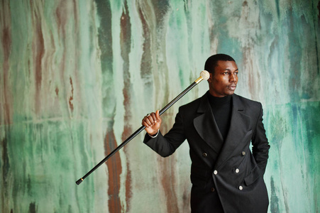 时尚的非裔美国人绅士，穿着优雅的黑色夹克，拿着复古的手杖，作为拐杖瓶或带金刚石球柄的鞭子。 富有时尚的非洲男人靠在绿色的墙上。