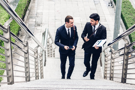 两个微笑的商人同事穿着黑色西装说话和走路。商人们在现代城市讨论战略