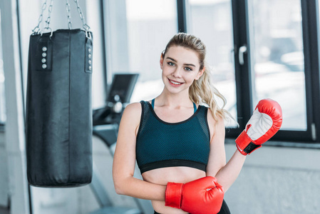 戴着拳击手套的快乐漂亮的运动女孩在健身房里对着镜头微笑