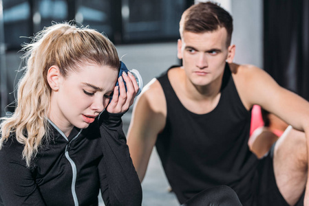 年轻的运动员看着心烦意乱的运动女孩把冰袋放在健身房的头上