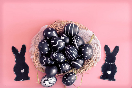复活节由黑色的鸡蛋组成，创造性的光图案手工制作，在一个由干草和复活节兔子制成的圆巢中，在粉红色的背景下分离出复活节假期的概念