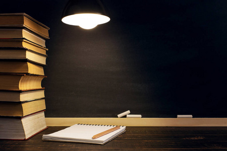 桌子背景下的粉笔板,书,笔记本和笔,在黑暗中的灯光下.复制空间.照片