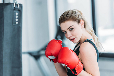 漂亮的年轻女运动员戴着拳击手套，在健身房里看着摄像机