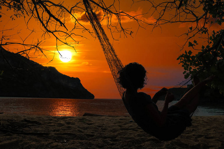 在日落和红天背景下，女人躺在海滩边的吊床上。