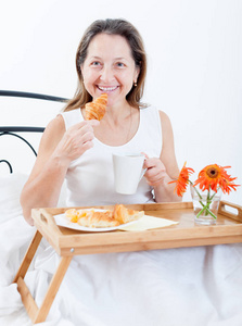 美丽的老妇人在床上卧室里吃早餐。