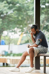 运动胡子的印度男人，穿着运动服，坐在户外的长凳上，跑步后脉搏跳动，看着手表