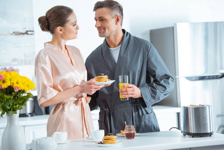 一对漂亮的夫妇在厨房吃早餐时穿着橙汁和煎饼的长袍