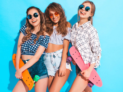 三个年轻时尚的微笑美丽的女孩与五颜六色的硬币滑板。 夏天的女人穿着时髦的格子衬衫，穿着太阳镜，摆在蓝墙附近的工作室里。 积极的模