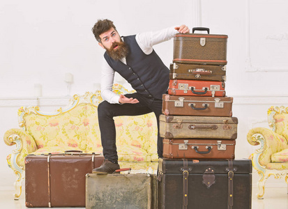 男士, 有胡子和胡须的管家提供行李, 奢华的白色内饰背景。男子气概优雅, 在震惊的脸上站在一堆老式手提箱。行李和旅行概念