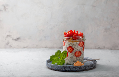 健康的早餐。 在灰色背景下的玻璃罐子里放着草莓辣椒种子的隔夜燕麦片