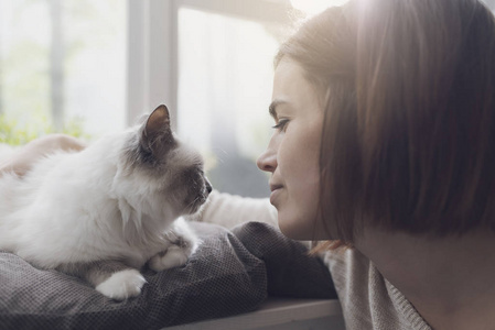 年轻女子在家中抚摸她漂亮的猫，旁边是窗户宠物和生活方式的概念