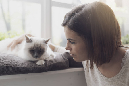 年轻女子在家中抚摸她漂亮的猫，旁边是窗户宠物和生活方式的概念