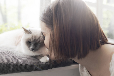 年轻女子在家中抚摸她漂亮的猫，旁边是窗户宠物和生活方式的概念