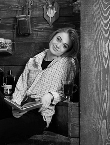 放松概念。女孩在休闲装坐与书在木古董内部。女孩的学生放松与书和玻璃的品酒。穿格子衣服笑脸的女士看起来很随意