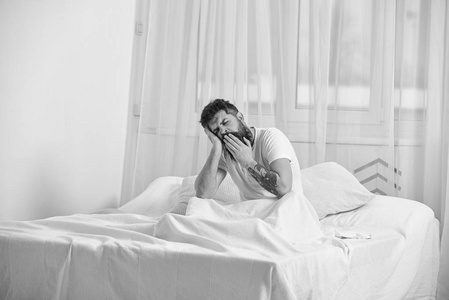 睡累了的人脸上打呵欠。穿衬衣的男人坐在床上, 背景上是白色的窗帘。男子气概的胡子和胡子打呵欠, 放松, 小睡, 休息。午睡和午睡