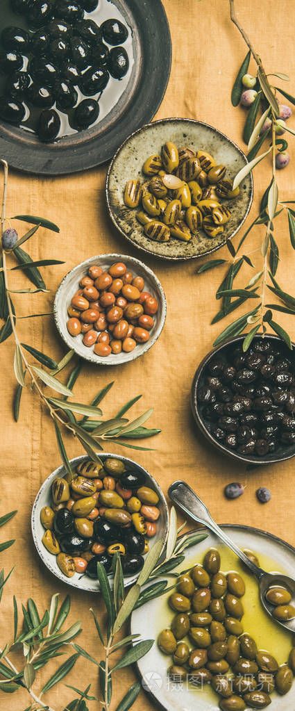 平铺各种地中海腌制橄榄在盘子和碗和橄榄树枝上的黄色亚麻背景，顶部视图。地中海美食开胃菜