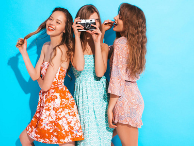三个年轻美丽的微笑时髦女孩穿着时髦的夏季休闲服装。无忧无虑的女人在蓝墙附近摆姿势。拍摄复古相机的照片
