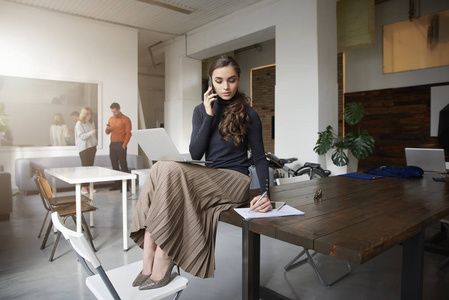 一个年轻的女商人坐在办公室工作时，用手机和别人说话。