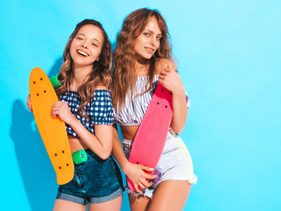 两个年轻时尚的微笑美丽的女孩与五颜六色的硬币滑板。 夏天的女人穿着时髦的格子衬衫衣服，摆在工作室蓝色墙壁附近。 积极的模特玩得开