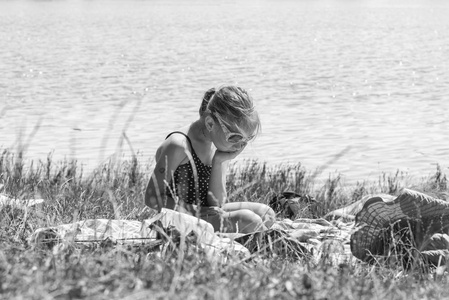 那个女孩在海滩上很伤心。 一个穿泳衣的女孩在阳光明媚的日子里孤独地坐在河边的草地上的海滩上。