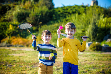 两个活跃的学龄前男孩在户外球场打羽毛球。 孩子们打网球。 儿童学校运动。 儿童运动员网拍和毽球运动。 朋友们一起快乐温暖的田野。