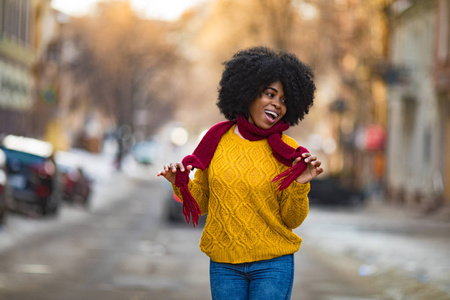 穿着围巾和毛衣的年轻黑人女人正对着城市街道笑。 特写镜头。