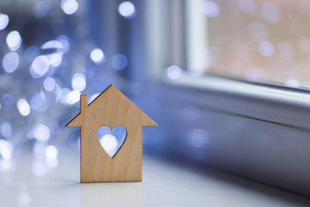 房子的木制图标，有洞的形式，在白天靠近窗户的心脏，在模糊的背景上有bokeh灯。带有复制空间的浪漫卡片。甜蜜的家的概念。