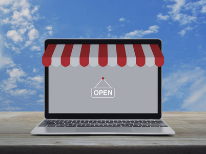 现代笔记本电脑，带有网上购物商店的图形和在蓝天上的木制桌子上的开放标志，带有白云商业互联网商店在线概念