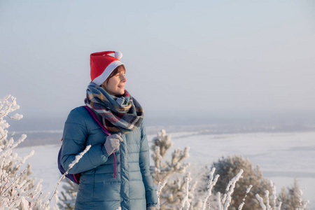 一个戴着红色圣诞帽的年轻女孩站在河岸上。 霜雪的冬日