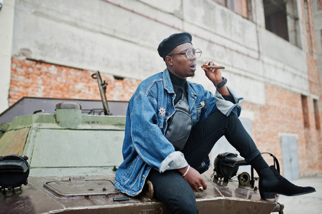 非裔美国人穿着牛仔裤夹克,贝雷帽和眼镜,抽着雪茄,对着btr军用装甲车