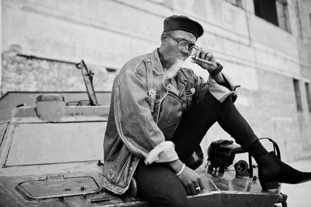 非裔美国人穿着牛仔裤夹克,贝雷帽和眼镜,抽着雪茄,对着btr军用装甲车