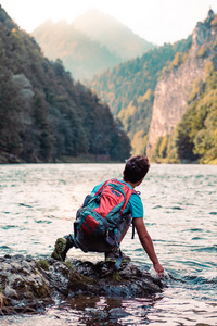 小男孩背着背包坐在河边的岩石上，手牵着水看着山峰，他穿着运动夏装
