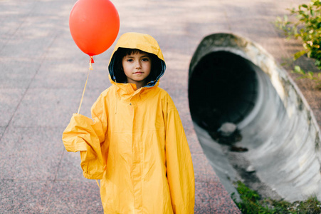 小女孩穿着超大号黄色雨衣,带着红色气球,靠在污水隧道管上照片