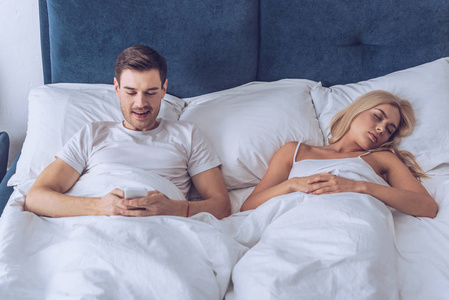 微笑的年轻人躺在床上，用智能手机，而年轻的妻子却睡着秘密的概念