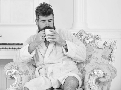男子睡浴袍, 喝咖啡, 享受豪华酒店的香气, 在早晨, 白色背景。胡子和胡须的男人坐在豪华扶手椅上享受早晨。精英休闲理念