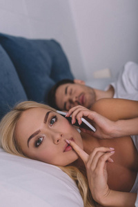 年轻女性用智能手机说话，用手指在嘴唇上看相机，而男朋友睡在床上的秘密概念