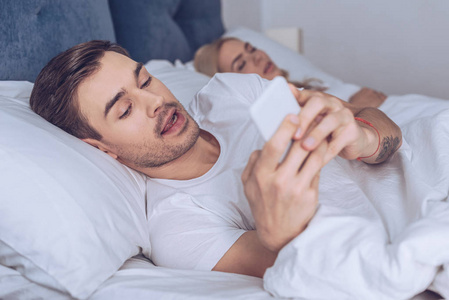 一个多疑的年轻人在和睡着的妻子躺在床上时使用智能手机