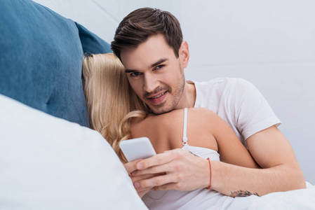 年轻人用智能手机，和女朋友躺在床上对着镜头微笑