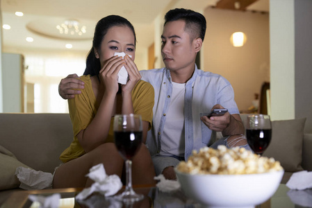 年轻的亚洲夫妇在看悲伤的电影，男人在沙发上安慰哭泣的女人