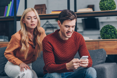 嫉妒的年轻女人看着微笑的丈夫在沙发上使用智能手机不信任的概念