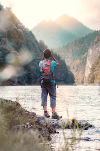 年轻的游客带着背包，通过望远镜在山顶上看，站在河上的岩石上。 男孩在山上度假，背着背包，他穿着运动的夏装