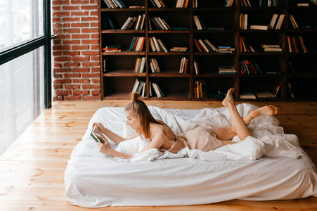 穿丝绸睡衣的漂亮女孩躺在床上，带着书在家