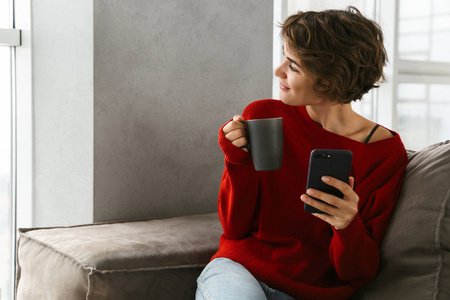 微笑的年轻女子穿着毛衣，在家里的沙发上放松，用手机喝咖啡。