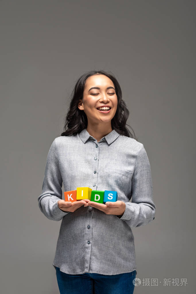 微笑着怀孕的亚洲年轻女子，手里拿着五颜六色的字母表立方体，把单词孩子隔离在灰色上