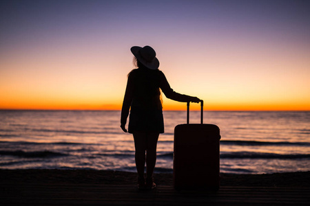 放松妇女与手提箱在海滩上日落剪影。假日旅行的概念。在海洋风景背景与手提箱的年轻夫人