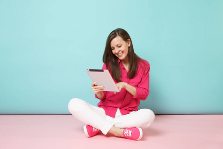 全长肖像女士，穿着玫瑰衬衫，衬衫，白色裤子，坐在地板上，手持平板电脑，孤立在粉红色的蓝色粉彩墙背景工作室。 时尚生活方式的概念。