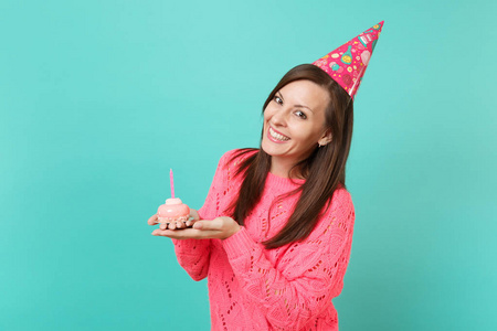 微笑的年轻女子穿着针织粉色毛衣和生日帽，手拿蛋糕，蜡烛隔离在蓝色绿松石墙背景工作室肖像上。 人们的生活方式概念。 模拟复制空间