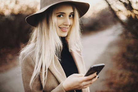 美丽的年轻女性，戴着时尚的帽子，微笑着看着相机，一边浏览智能手机，站在秋天乡村模糊的背景上。微笑的女人在农村使用智能手机