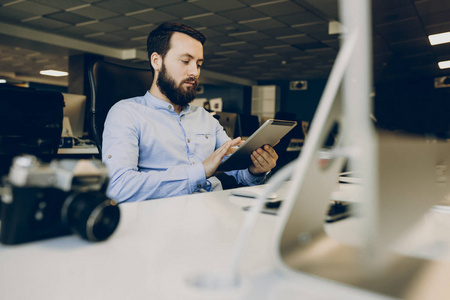 英俊的留胡子的家伙使用现代平板电脑，而坐在餐桌上的时尚办公室。办公室里使用平板电脑的胡子男