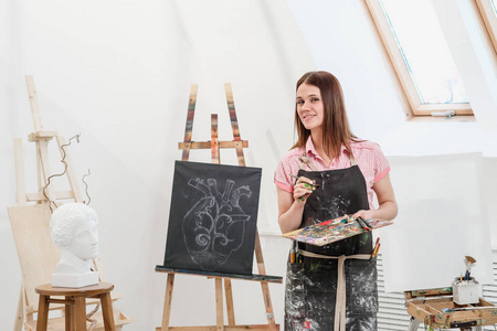 一位年轻的女画家在明亮的白色工作室里在画架上的画布上画一幅画